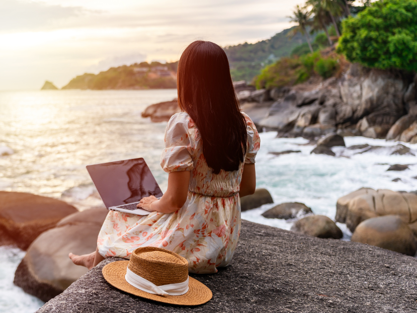 Woman sitting on beach rocks with laptop and straw hat overlooking the ocean