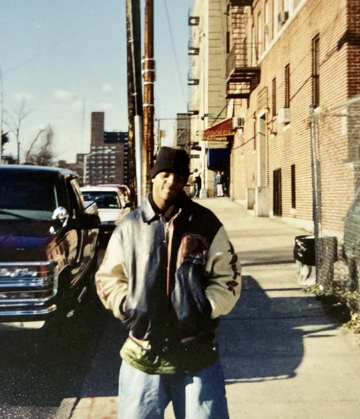 Young man in varsity jacket and beanie standing on a city sidewalk with brick buildings