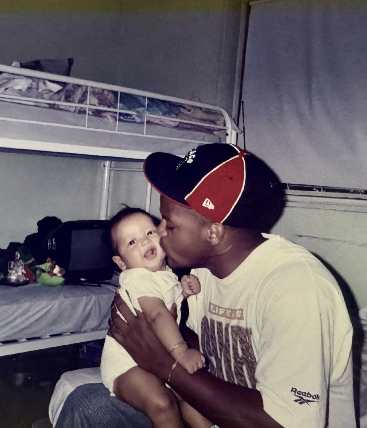 Man in baseball cap holding a smiling baby in a room with bunk beds