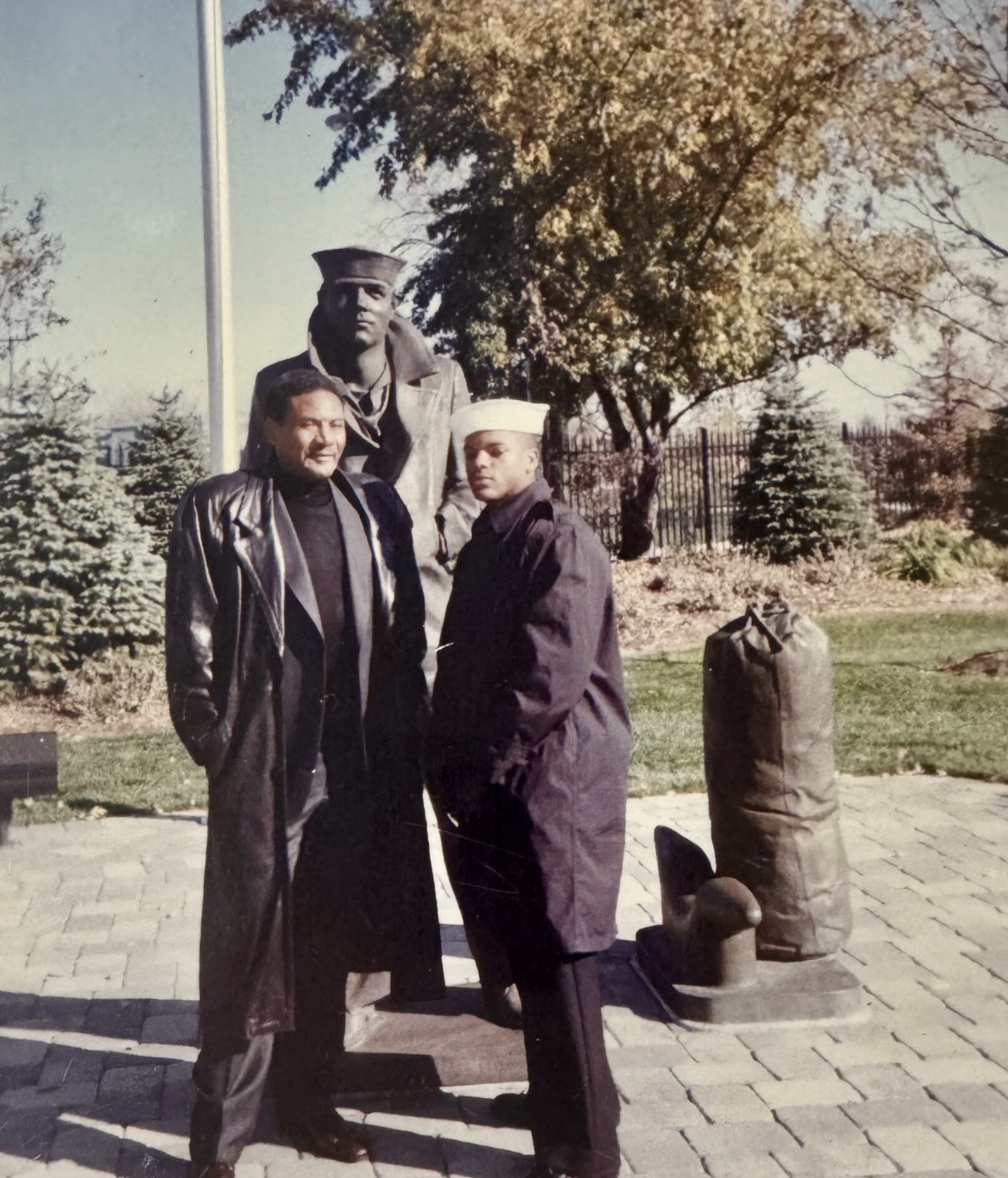 Two men in U.S. Navy uniforms standing beside a naval statue outdoors