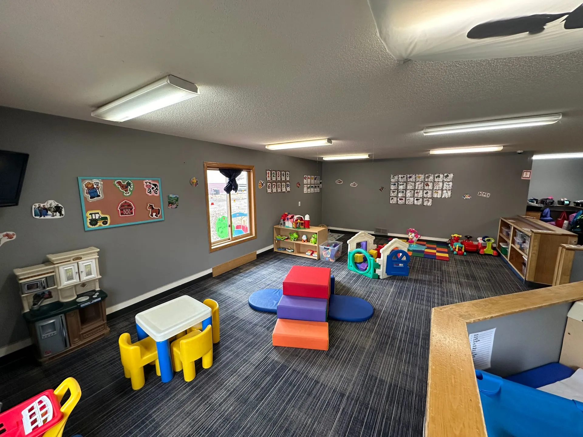 Bright toddler classroom with soft climbing blocks, play kitchen, and small activity tables