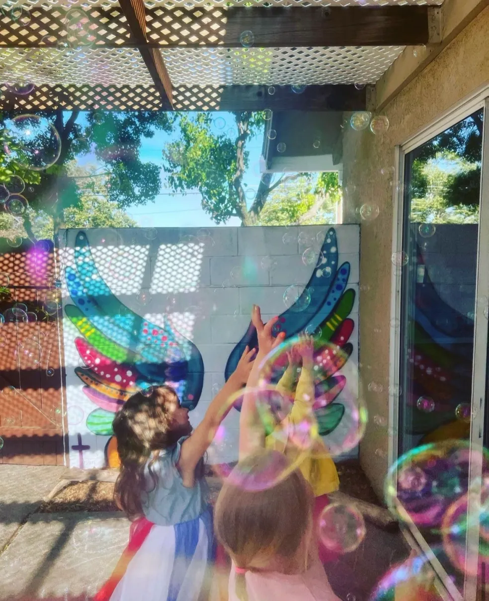 Children laughing and chasing bubbles against the colorful angel wings mural