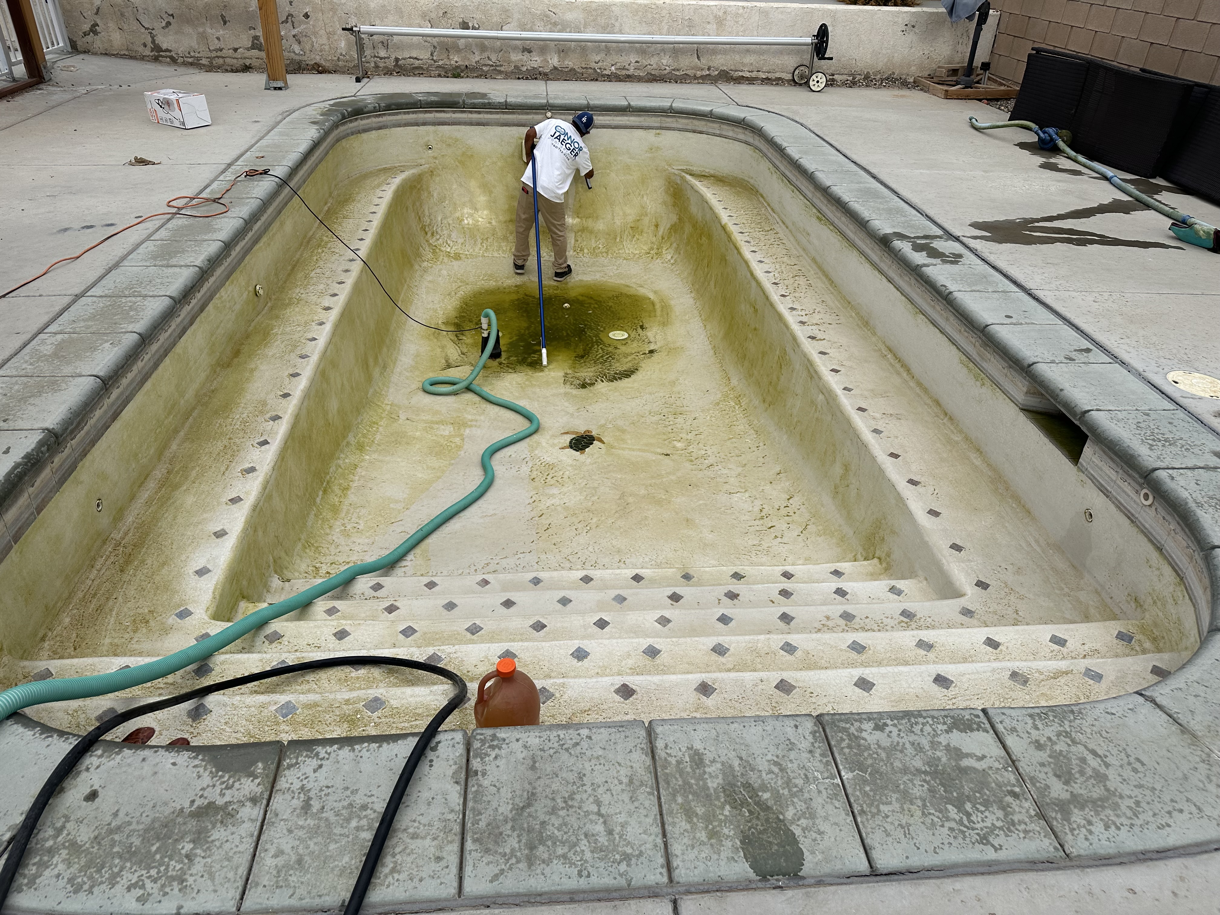 Drained pool with heavy yellow algae, mineral stains, and dirt being scrubbed by Connor Jaeger in Ridgecrest, CA