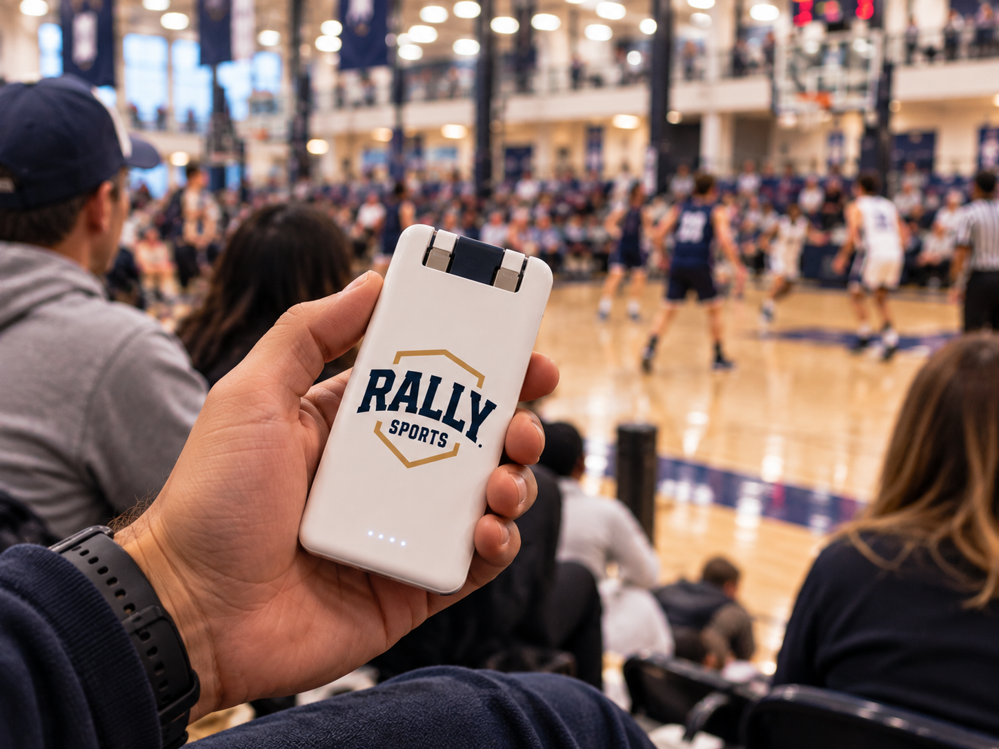 A spectator seated in the bleachers holding a RALLY SPORTS branded portable battery while using her phone during a basketball game