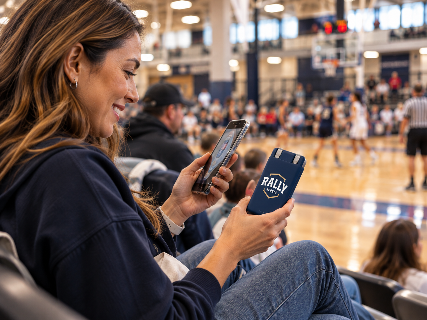 A spectator seated in the bleachers holding a RALLY SPORTS branded portable battery while using her phone during a basketball game