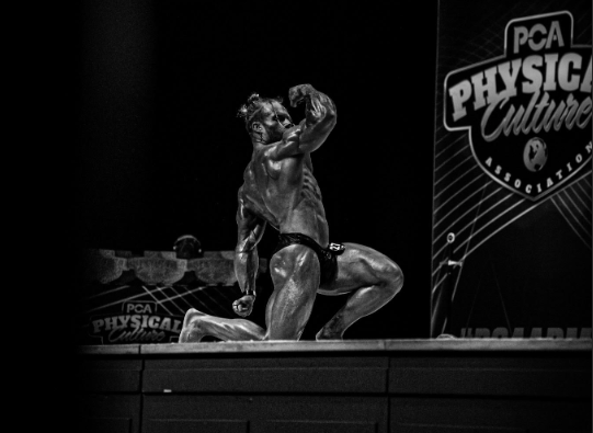 James Alabaster PCA onstage kneeling back and arm pose showing muscular conditioning