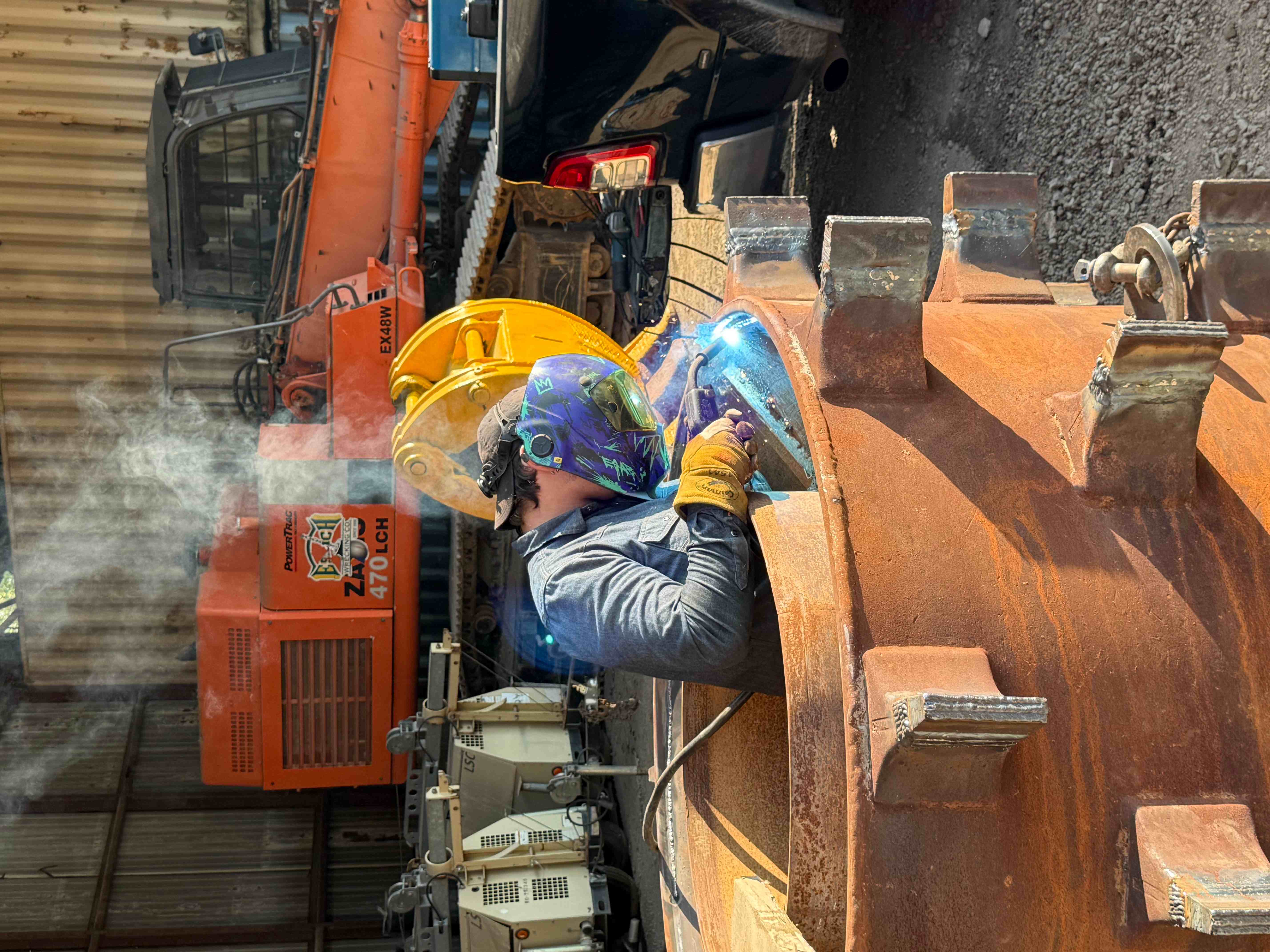 Welder in full hood welding wear blocks onto a heavy steel shell at an equipment yard