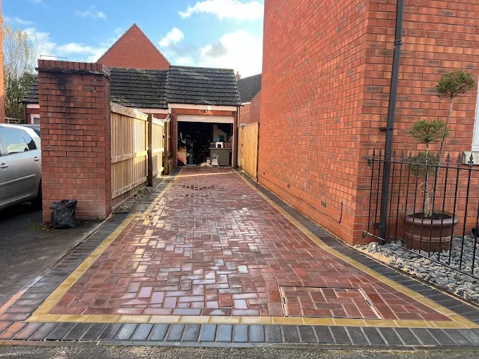 Long brick block paving driveway leading to detached garage with recessed manhole cover and dual-tone border