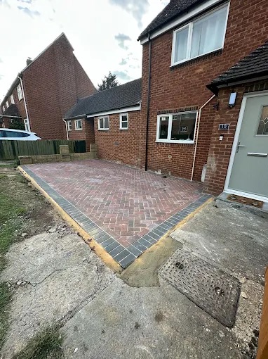 Red-tone herringbone block paving parking bay with charcoal kerb border outside brick home