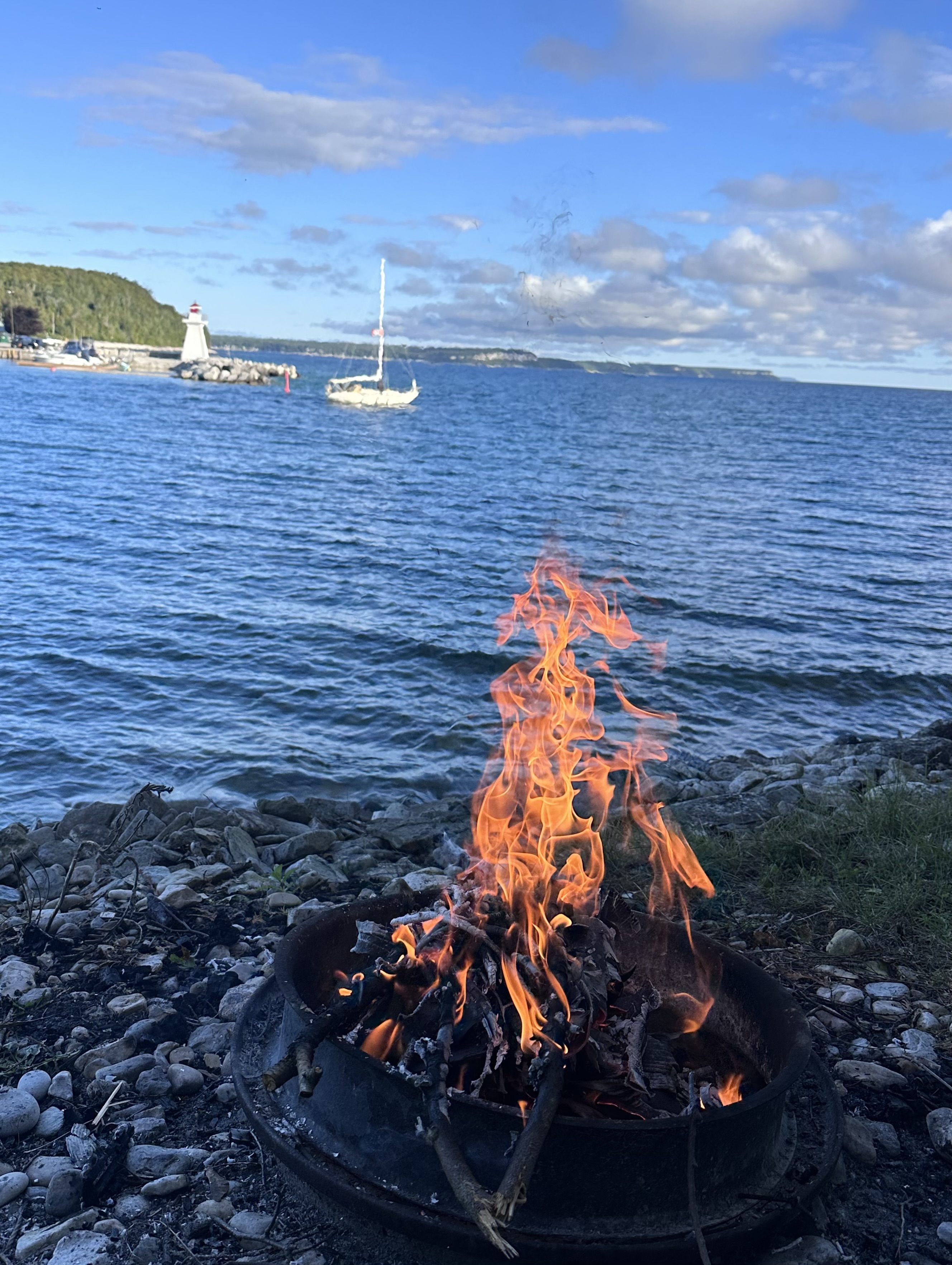 Shoreline campfire flames overlooking Georgian Bay with passing sailboat and lighthouse
