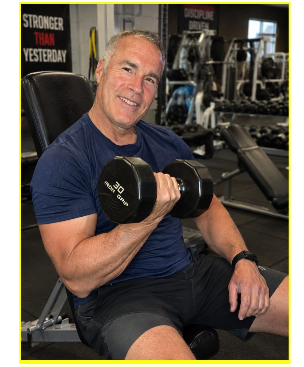 A strong, smiling man over 40 seated on a gym bench curling a 30-pound dumbbell — progressive resistance training stimulating muscle protein synthesis