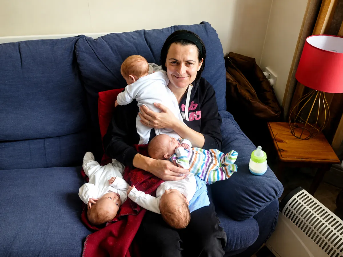 Lalena smiling on a blue armchair holding and surrounded by her newborn quadruplets