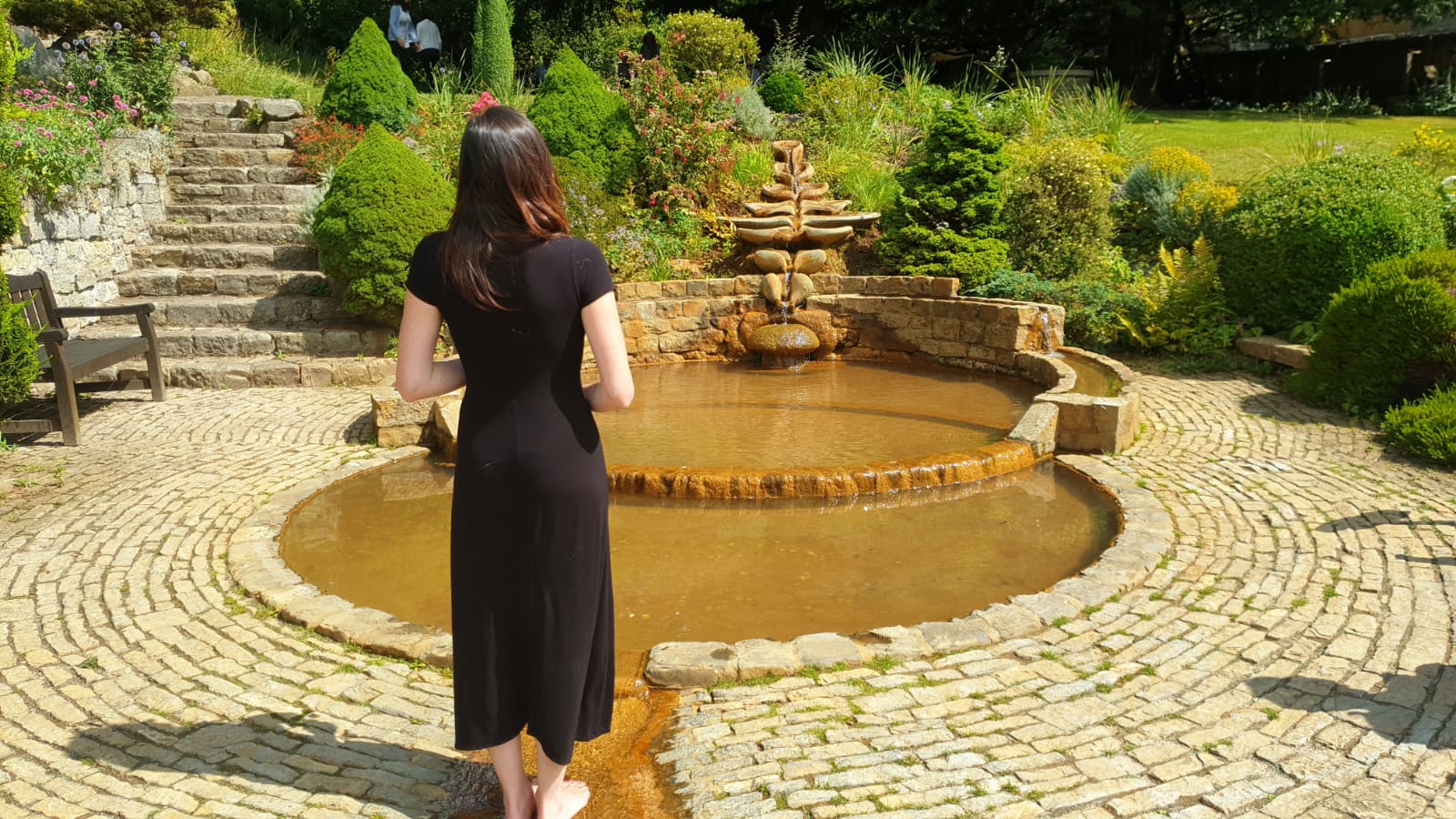 Lalena Rose in a black dress standing barefoot by a stone water garden with cascading flowforms