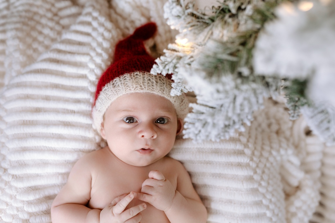 Wide-eyed baby wearing red Santa cap next to frosted Christmas tree