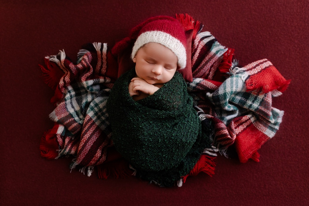 Peaceful sleeping baby wrapped in pine green with red Santa hat on burgundy backdrop