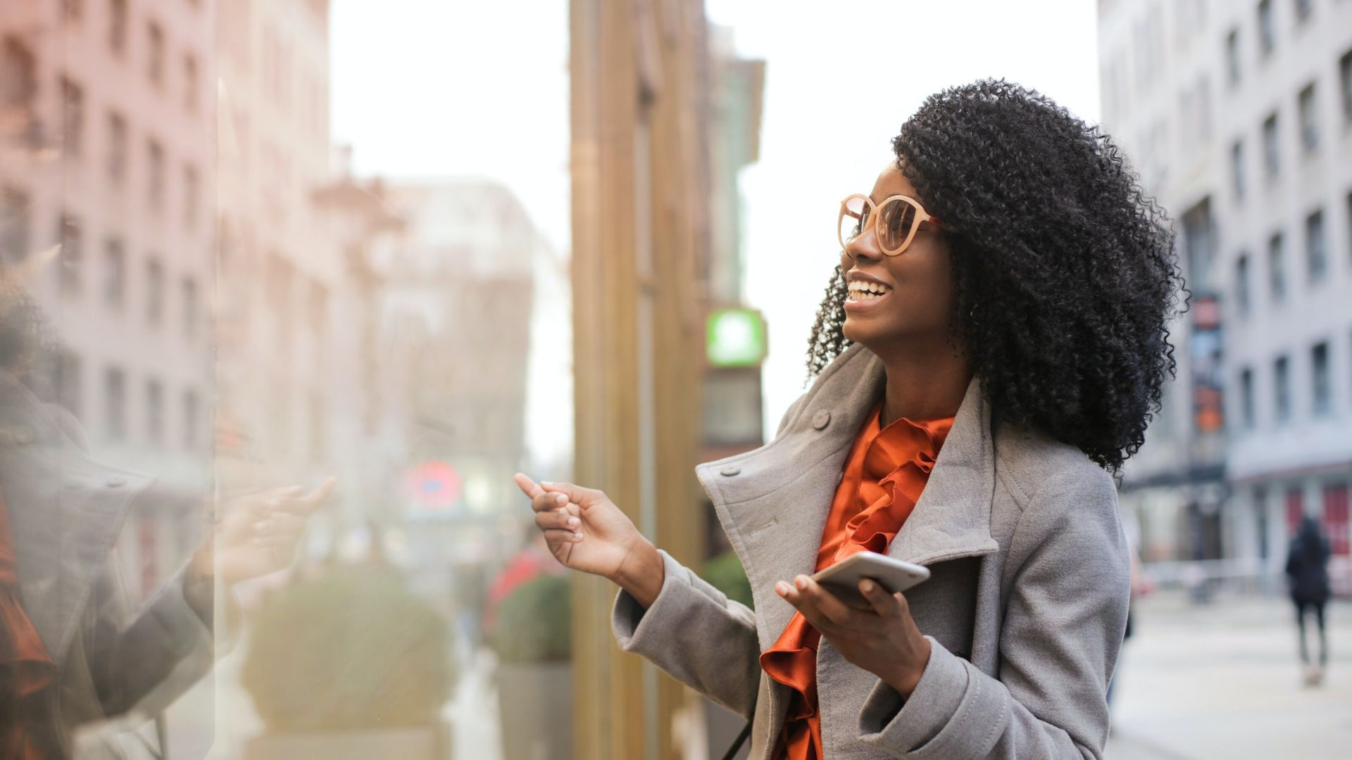 Woman smiling radiantly looking into shop window with phone in hand