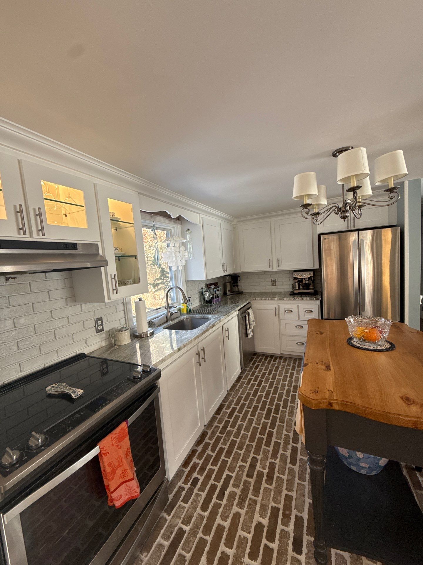 Completed white painted kitchen cabinets with interior illuminated glass cabinets, brushed nickel pulls, and rustic brick floor