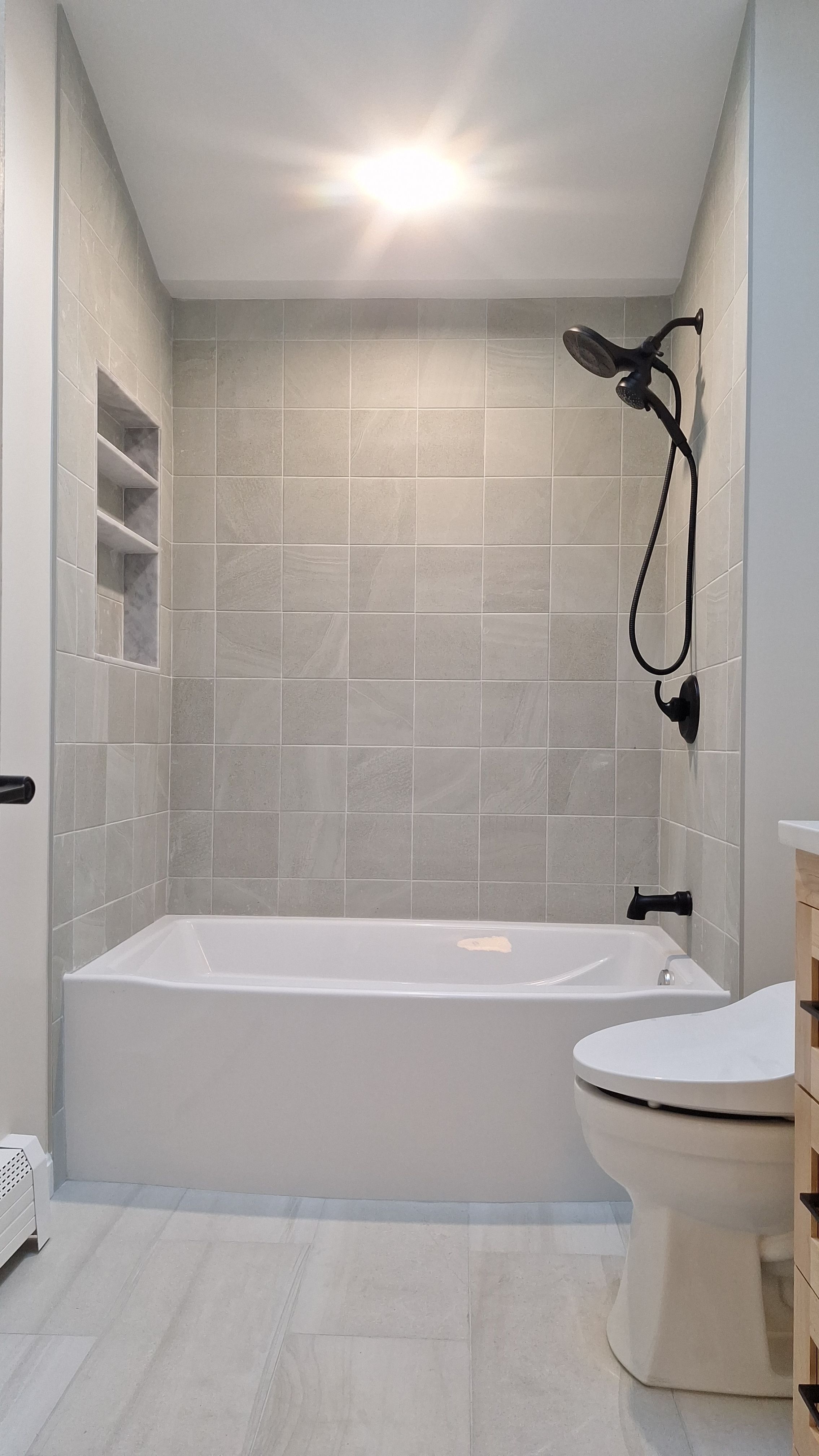 Completed bathroom remodel with clean white soaking tub, soft grey tile surround, recessed marble shelf niche, and matte black fixtures