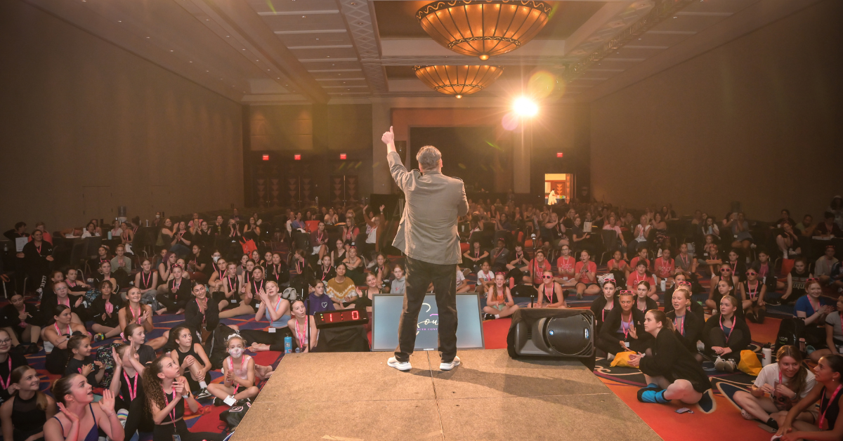 Rhee Gold on stage with his back to the camera giving a thumbs-up to a large energized ballroom of dance students and educators
