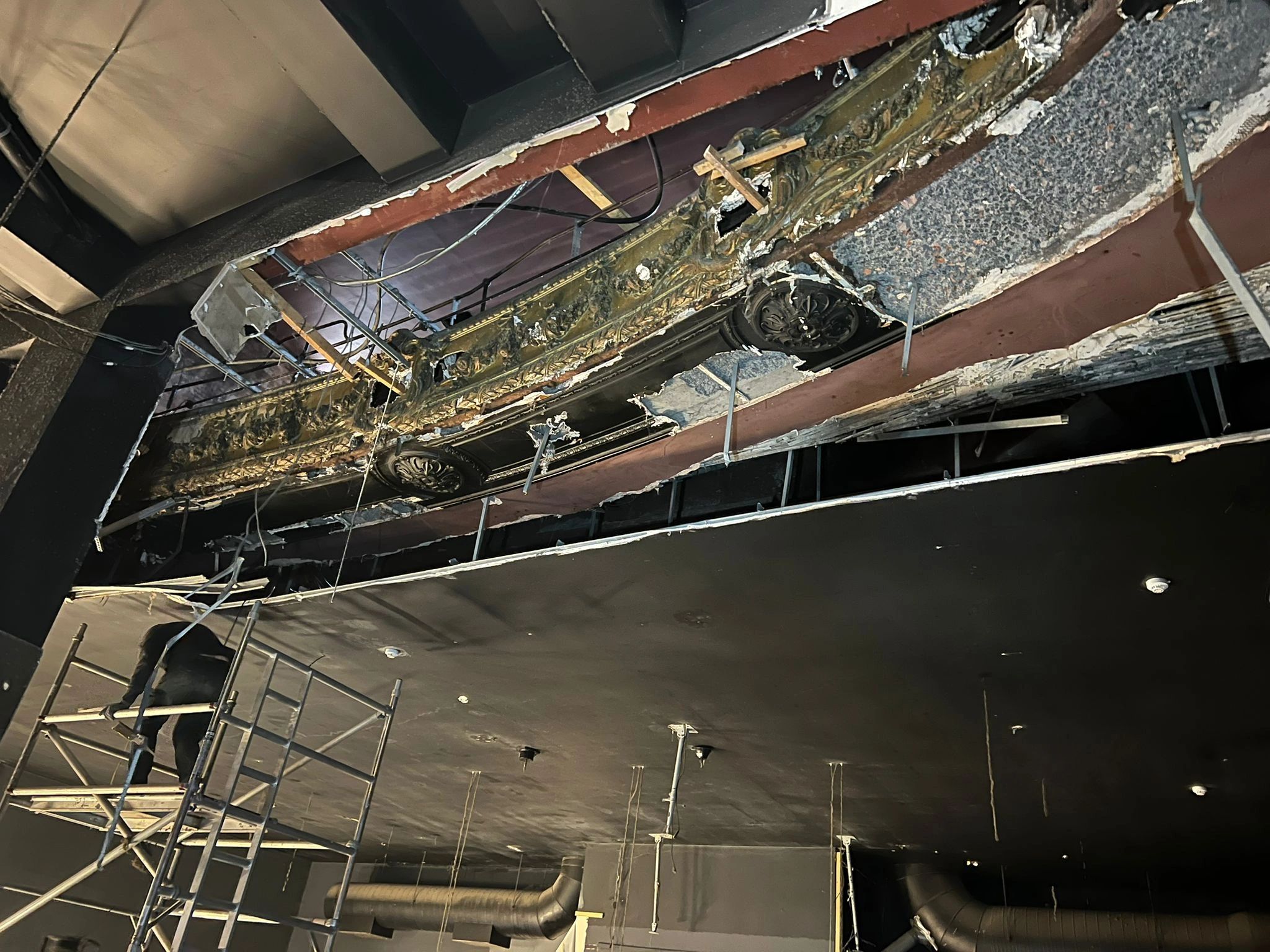 A worker on scaffolding repairs a ceiling with exposed beams and insulation.