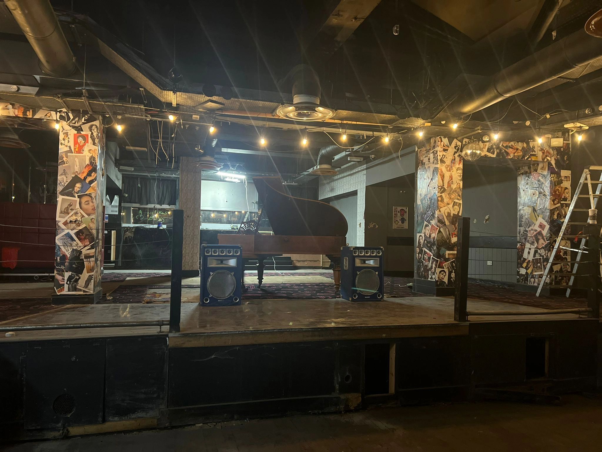 Dimly lit stage with a grand piano and amplifiers under string lights.