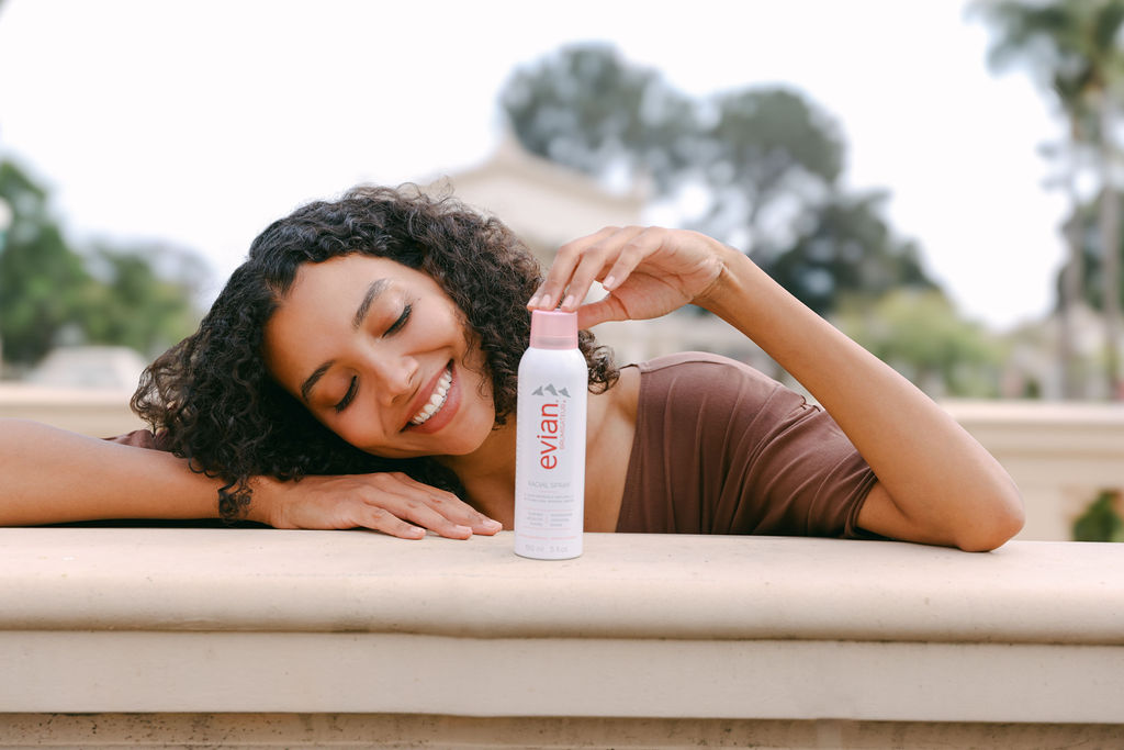 Campaign photography demonstrating product use — smiling woman holding a facial mist spray outdoors in natural light.