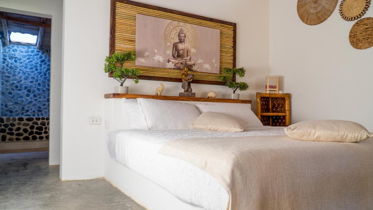 White linen bed with Buddha artwork on bamboo, bedside bonsai, and doorway into private stone bathroom at Bali Lodge