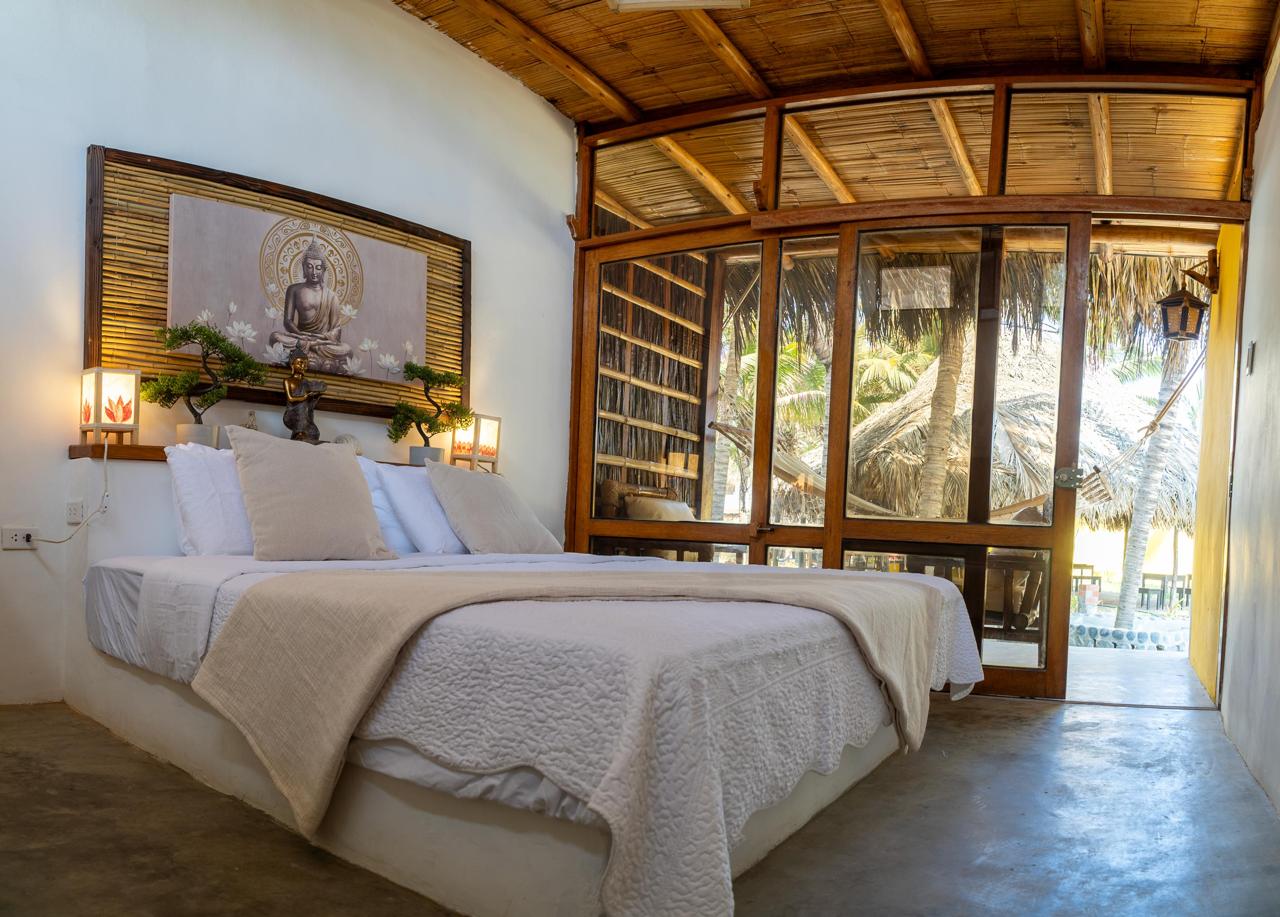 Uluwatu Suite at Bali Lodge Hotel — double bed with Buddha artwork, bamboo slats, warm lantern lamps, and timber glass doors opening to palapa terrace
