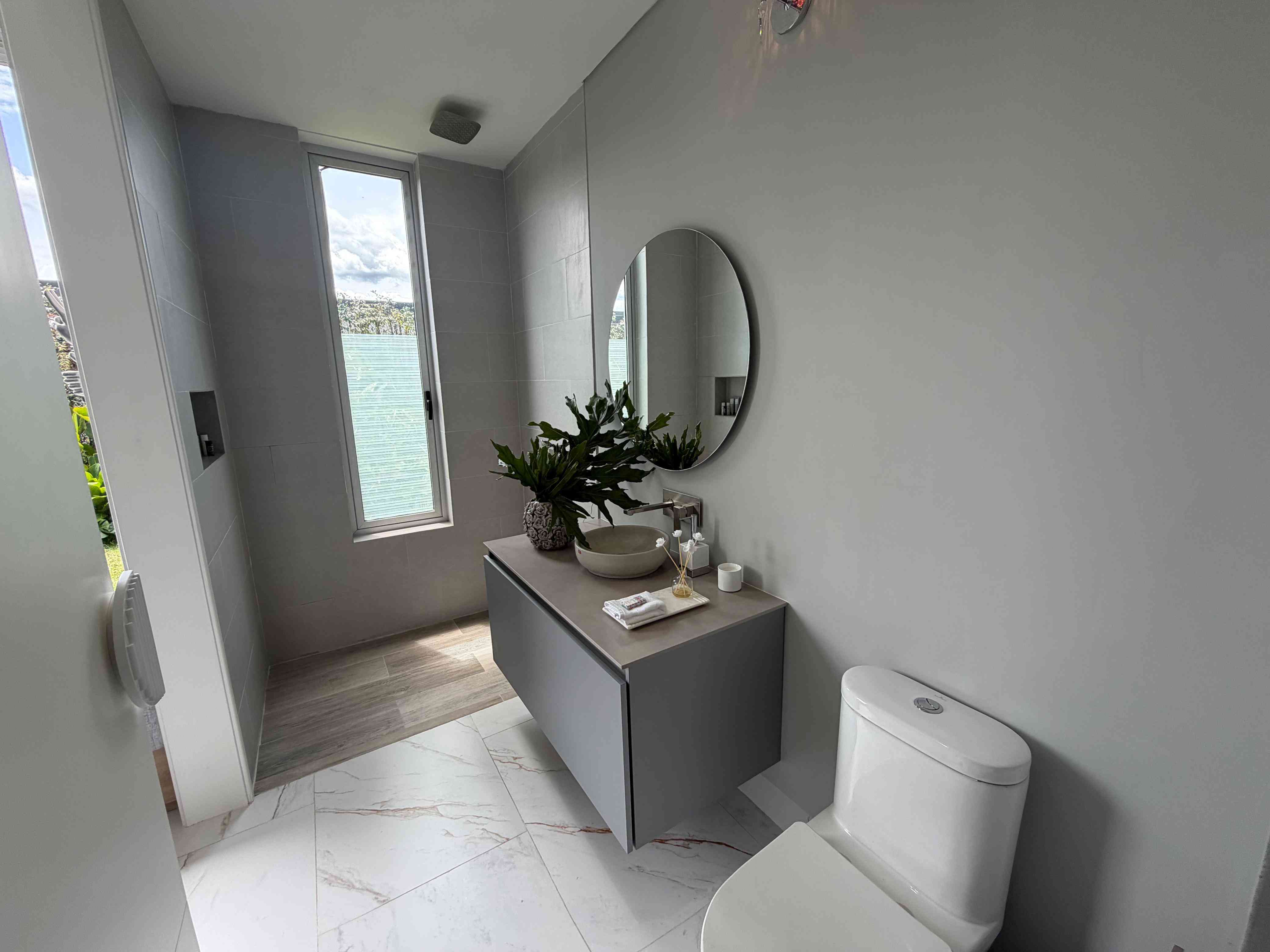 Baño auxiliar moderno con espejo circular, lavamanos de diseño y ventana natural