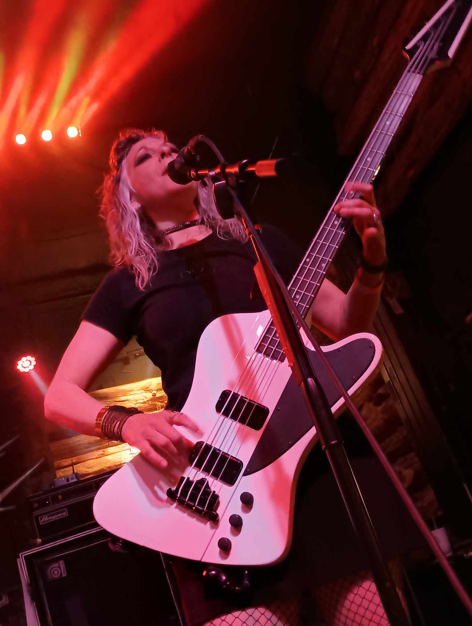 Johnnie Kay singing and playing white bass guitar live on stage under red stage lighting