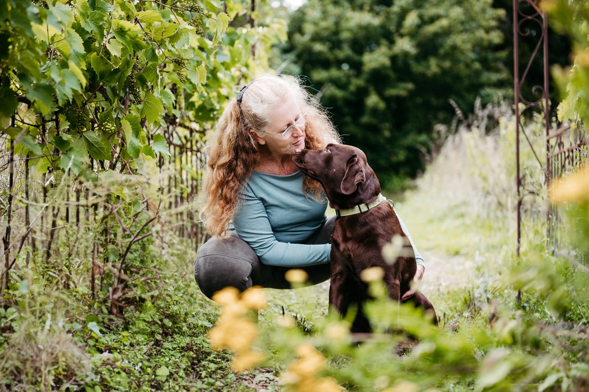 Dr. Viviane Theby hockt entspannt im Grünen mit ihrem schokoladenbraunen Hund, der ihr zugewandt ist
