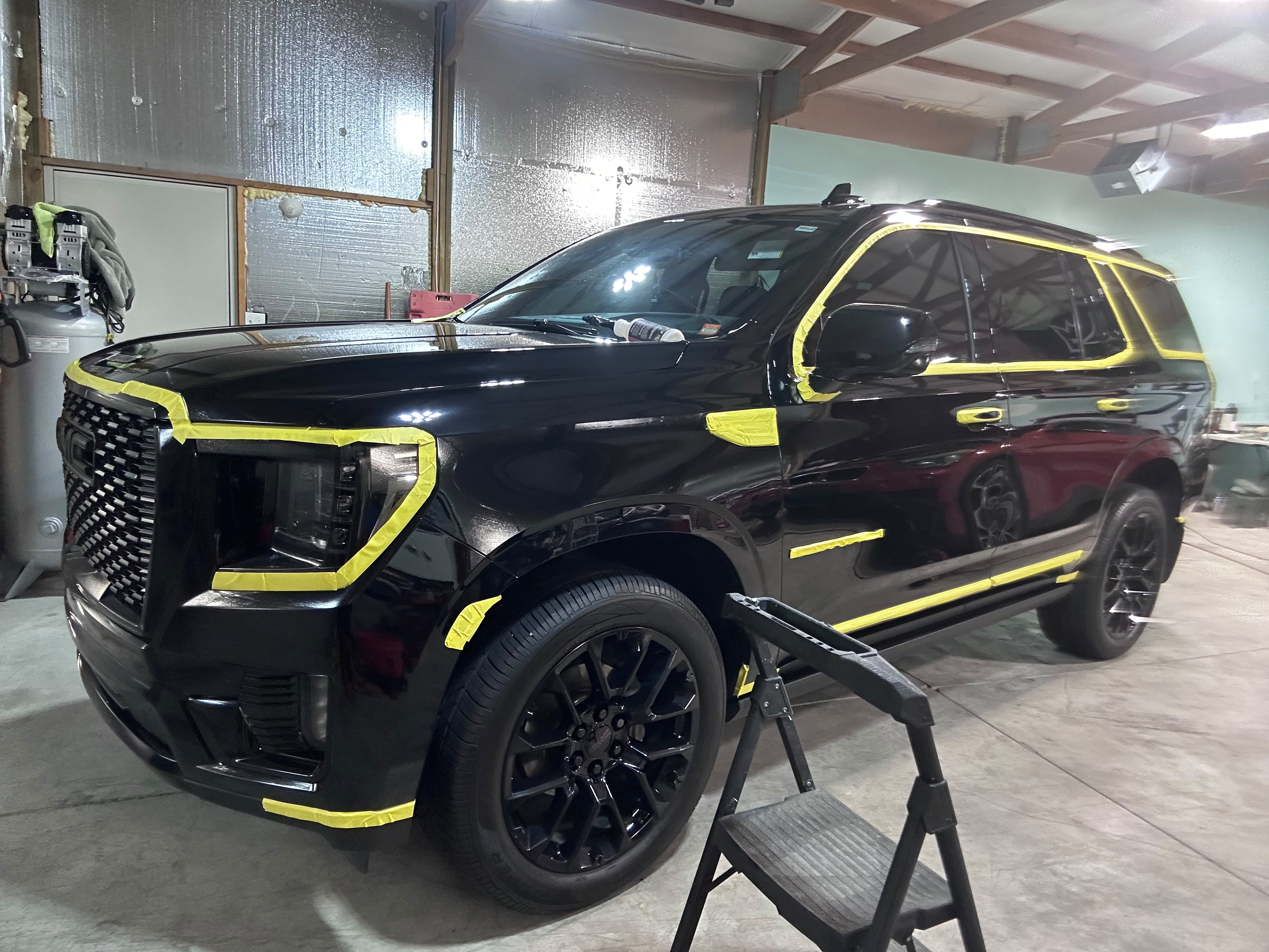 Black GMC Yukon Denali masked with yellow detailing tape across trims, emblems, and body lines for multi-stage compounding polish in Miracle Detailing shop bay