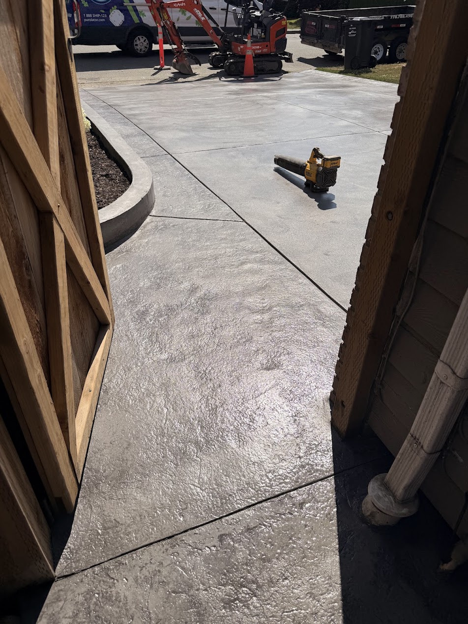 View through a wooden gate of textured concrete, control joints and a curved raised edge, with a tool on the surface.