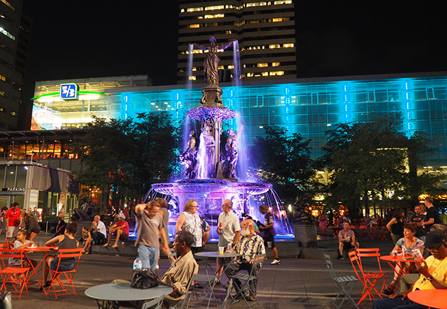 The Tyler Davidson Fountain illuminated at night on Fountain Square with people gathered at outdoor café tables in Downtown Cincinnati