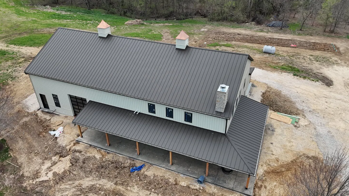 Aerial view of a large barndominium with a bronze standing seam metal roof, copper topped cupolas and a wraparound porch roof, installed by Snaplock Roofing LLC