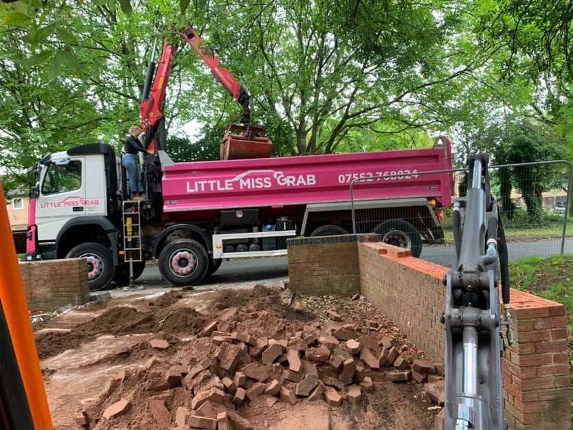 Operator using hydraulic grab crane arm to scoop hardcore and rubble over garden wall