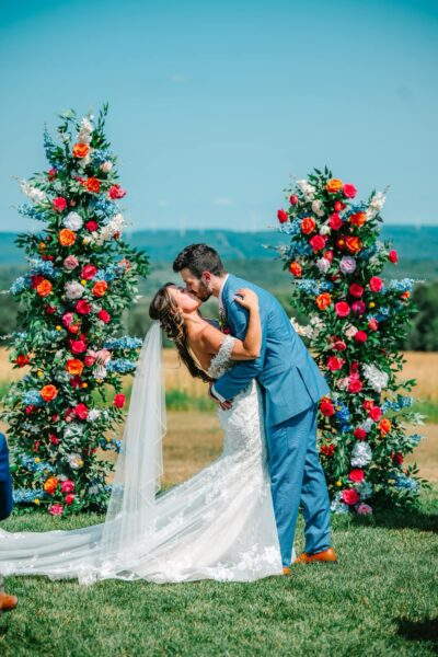 Bride and groom sharing a dip kiss between colorful floral pillars under bright summer blue sky