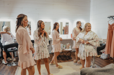 Bride and bridesmaids in blush robes gathering in the bridal dressing room with illuminated vanity mirrors