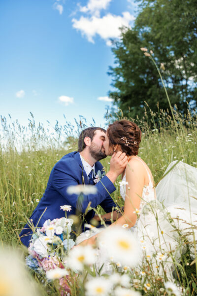 Bride and groom kissing amidst lush blooming wild daisies and green meadows under a blue spring sky
