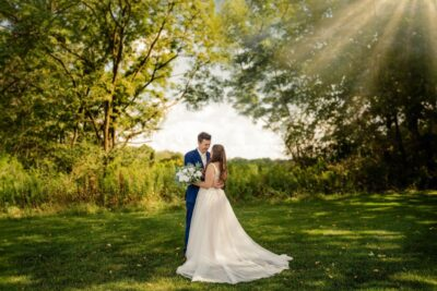 Bride and groom embracing in lush green woodland meadow with sunbeams filtering through trees