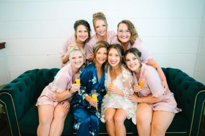Bride and bridesmaids in satin blush robes raising champagne glasses together on the bridal suite sofa