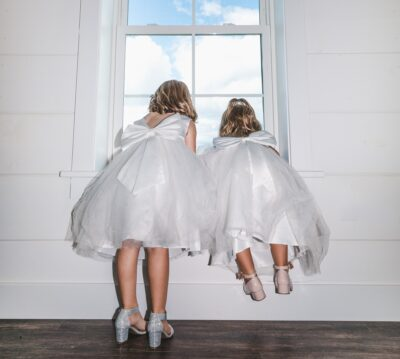Two young flower girls in white tulle dresses peeking through window at blue sky
