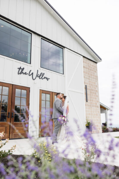 Bride and groom sharing a kiss in front of The Willow barn doors framed by blooming purple spring flowers