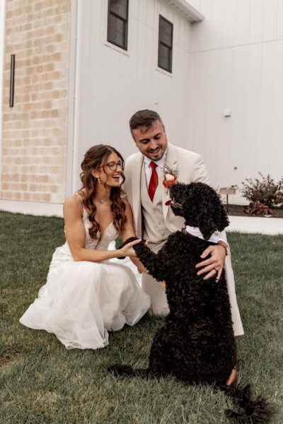 Bride and groom kneeling on the lawn laughing and holding paws with their beloved black doodle dog