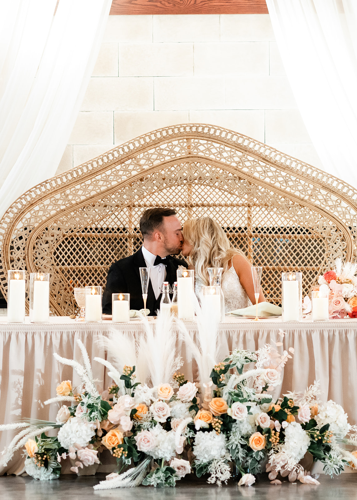 Bride and groom kissing at elegant sweetheart table with wicker peacock chair, candles, and floral installation