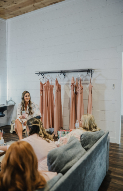 Bride smiling in bridal suite with blush bridesmaid dresses hanging along the white shiplap wall