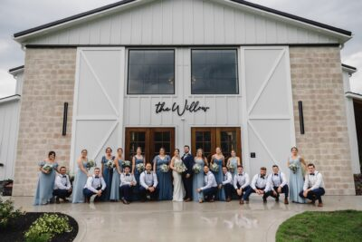 Full wedding party in dusty blue dresses and suits lined up in front of The Willow venue building