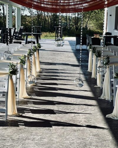 Covered patio ceremony aisle lined with white chairs, hanging glass terrariums, and fabric draping