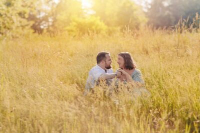 Couple sitting close and smiling together in golden meadow grass during warm sunset golden hour