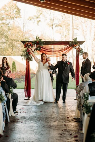 Bride and groom raising hands celebrating down the ceremony aisle with burgundy draping